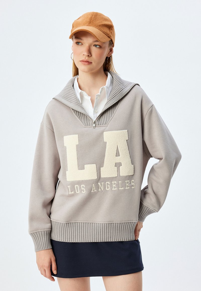 Jasnoszary sweatshirt z dużym białym napisem "LA", prążkowanym kołnierzem i mankietami, z zamkiem błyskawicznym przy szyi, noszony z białą koszulą i granatową spódnicą.