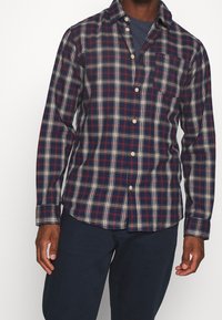 Camicia a quadri in blu navy, rosso e bianco; maniche lunghe, chiusura frontale con bottoni, taschino sul petto singolo, tessuto misto cotone, vestibilità comoda.