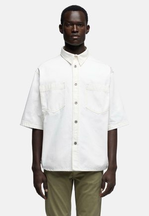 Man wearing a loose white button-up shirt with short sleeves and large chest pockets, paired with olive green pants, facing forward.