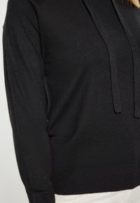 Felpa nera con cappuccio dal tocco morbido, dotata di una tasca frontale e coulisse. Il design è a vestibilità comoda con bordi a costine.