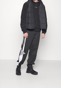 Gilet rembourré noir isolé par-dessus un sweat à capuche noir, associé à un pantalon de survêtement gris avec des panneaux latéraux blancs et gris. Bottes noires à lacets.