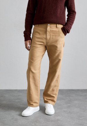 Person som bär beige raka byxor, mörkröd tröja och vita sneakers, stående på grå betonggolv mot vit vägg.