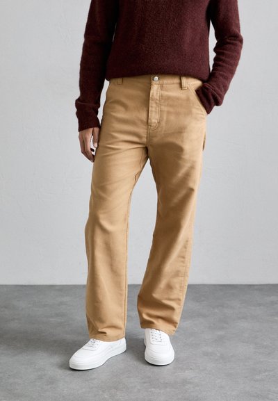 Persona in pantaloni beige a gamba dritta, maglione rosso scuro e sneakers bianche, in piedi su un pavimento in cemento grigio contro un muro bianco.