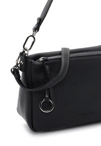 Sac bandoulière en cuir noir texturé avec fermeture éclair argentée, anneau métallique, sangle ajustable et nom de la marque "Suri Frey" discrètement embossé.