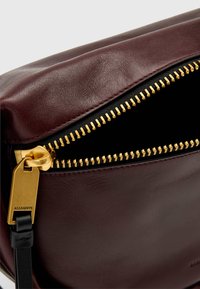 Bordeaux lærveske med gullglidelås og beslag. Har en glatt tekstur og svart interiør, og viser detaljerte sømmer langs kanten.