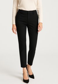 Pantaloni neri su misura con una vestibilità slim, caratterizzati da una texture liscia e passanti per la cintura, abbinati a tacchi a punta neri e a una blusa di un leggero colore crema.