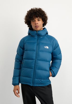 Giacca a vento blu con sezioni cucite, cappuccio e zip frontale completa. Presenta polsini elasticizzati e un logo sul lato sinistro del petto.