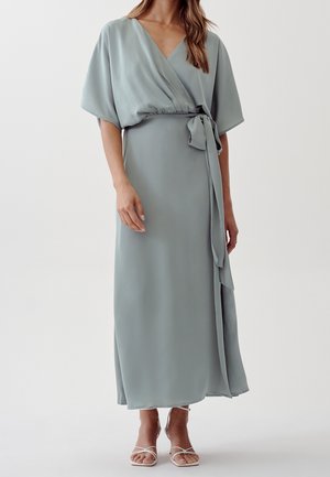 Kvinde iført en lyseblå slå-om-kjole med korte, brede ærmer, bundet talje og hvide højhælede sandaler med stropper mod en ensfarvet baggrund.