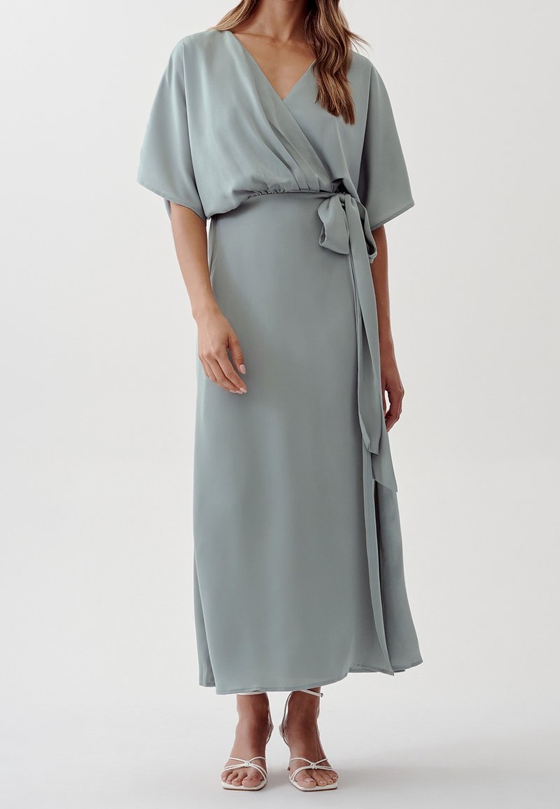 Femme portant une robe portefeuille bleu clair avec des manches courtes larges, une taille nouée, et des sandales blanches à talons hauts à lanières sur un fond uni.
