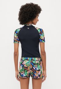 Czarny rash guard z krótkim rękawem i kolorowymi tropikalnymi rękawami, w połączeniu z wzorzystymi szortami w odcieniach zieleni, różu i brązu.