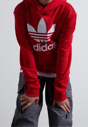 Person iført en rød Adidas-hoodie og grå cargobukser, bøjet fremad med hænderne hvilende på lårene mod en neutral baggrund.