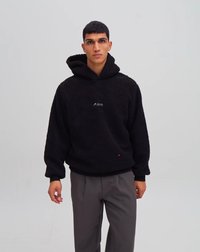 Crni dukserica s kapuljačom od flisa s teksturiranom vanjštinom, koja sadrži mali logo na prsima i crveni naglasak na rubu. Model nosi sive hlače.