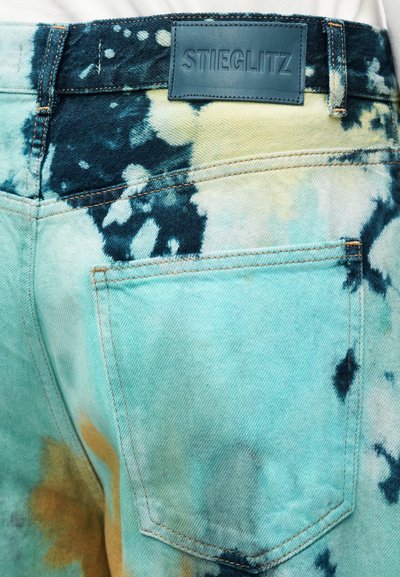 Τσέπη πίσω και ζώνη της μέσης σε τζιν με σχέδιο tie-dye σε μπλε, κίτρινο και μαύρο, με μια μαύρη ετικέτα της μάρκας Stieglitz στη ζώνη της μέσης.