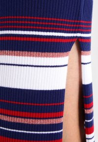 Dettaglio di un maglione lavorato a maglia con strisce blu navy, rosse e bianche e una texture a costine. Presenta un'apertura laterale per un design aggiuntivo.
