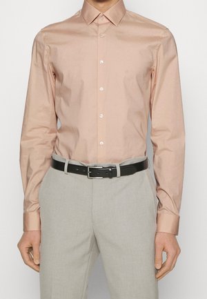 Man wearing a pale pink button-up shirt with long sleeves, tucked into light grey trousers secured with a black belt.