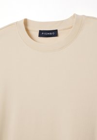 T-shirt in cotone beige con collo rotondo e maniche corte. L'etichetta riporta la scritta "PIOMBO" su un'etichetta di colore scuro a contrasto. Texture liscia.