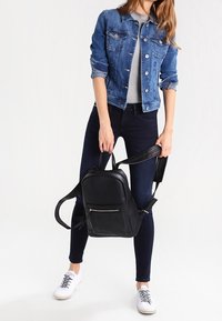 Svart läderryggsäck med texturerad yta, som har en framficka med dragkedja, justerbara axelremmar och rundad form. Bärs av en person.