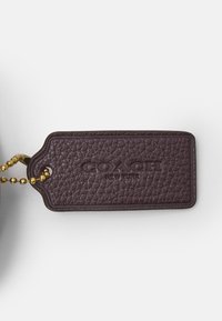 Brązowa skórzana metka o fakturowanej powierzchni, z wytłoczonym logo "COACH NEW YORK", przymocowana złotym łańcuszkiem z koralikami.