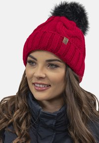 Cappello beanie rosso lavorato a maglia con dettagli a treccia e un pompon in pelle ecologica nera. Presenta un piccolo etichetta marrone sul lato. Indossato con una giacca blu navy.