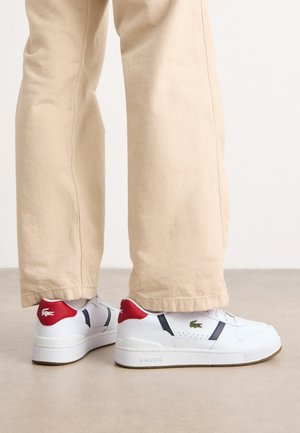 Personne portant un pantalon beige et des baskets blanches Lacoste avec des talons rouges et un logo crocodile vert, debout sur un sol clair.