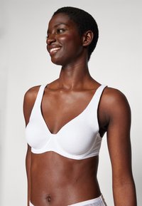 White underwire bra with textured fabric, deep V-neck design, wide straps, and smooth finish, worn by a model with short hair.