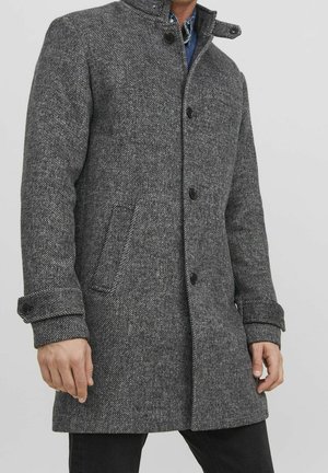 Homme portant un manteau en laine gris texturé avec des boutons et des poches avant sur un pantalon foncé, debout devant un fond clair uni.