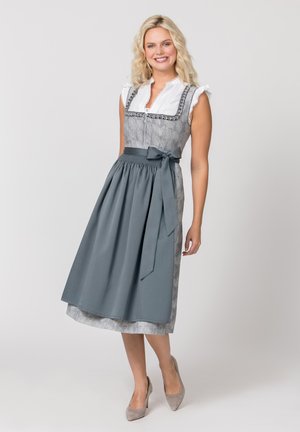 Junge Frau lächelt und trägt ein traditionelles graues Dirndl mit weißer Bluse und passender grauer Schürze und Absatzschuhen.