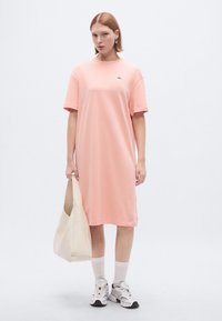 Ροζ ανοιχτό ροζ oversized φόρεμα T-shirt, κοντά μανίκια, στρογγυλός λαιμός, βαμβακερό υλικό, με λογότυπο στο στήθος, συνδυασμένο με κρεμ τσάντα ώμου και αθλητικά παπούτσια.