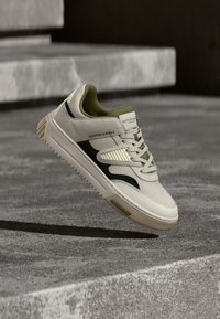 Weißes und olivgrünes Sneaker-Modell mit strukturierter Oberseite, schwarzen Akzenten und gepolstertem Kragen. Verfügt über eine Gummisohle und durchbrochene Designelemente.