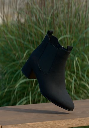 Bottine en daim noir à talon bloc sur un banc en bois, arrière-plan d’herbe haute verte, vue de côté avant.