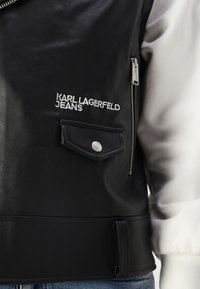 Black leather jacket with a silver zipper, featuring an embroidered logo, a flap pocket, and a textured finish. Includes a contrasting white sleeve.