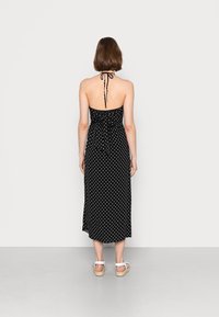 Schwarzes Kleid mit Polka-Dots, tailliertem Oberteil, Halternacken und Krawattenverschluss auf der Rückseite. Die Länge reicht bis zur Mitte der Waden und wird mit weißen, schmalen Sandalen kombiniert.