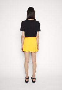 T-shirt noir à manches courtes associé à une mini-jupe plissée jaune, présentant une texture lisse et un ourlet évasé. Sandales noires complètent la tenue.