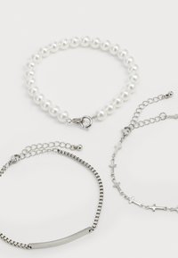Trois bracelets : l'un avec des perles de coquillage blanc et un fermoir en métal, l'autre avec une chaîne en argent et un détail en barre, et un autre avec de petits motifs de croix.