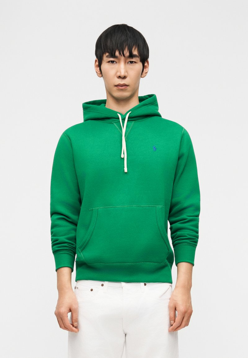 Mladi moški, oblečen v zeleno kapuco z belimi vrvmi in belimi hlačami, stoji ob svetlem enobarvnem ozadju.