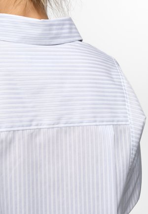 Nærbilde av en person som har på seg en hvit skjorte med tynne horisontale og vertikale lyseblå striper og oppbrettet krage.