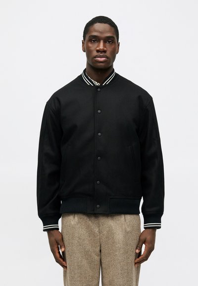Schwarze Collegejacke mit Druckknöpfen, gerippten Bündchen und Kragen, der mit weißen Streifen verziert ist; getragen über strukturierten braunen Hosen.