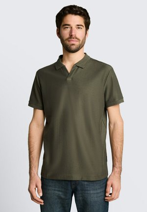 Mann mit kurzen dunklen Haaren und Bart, trägt ein olivgrünes kurzärmeliges Poloshirt und blaue Jeans, steht vor hellem Hintergrund.