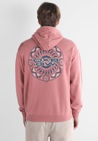 Felpa rosa con una grande grafica circolare blu scuro sul retro che presenta il testo "OXBOW" e motivi intricati. Tessuto di cotone, vestibilità comoda.