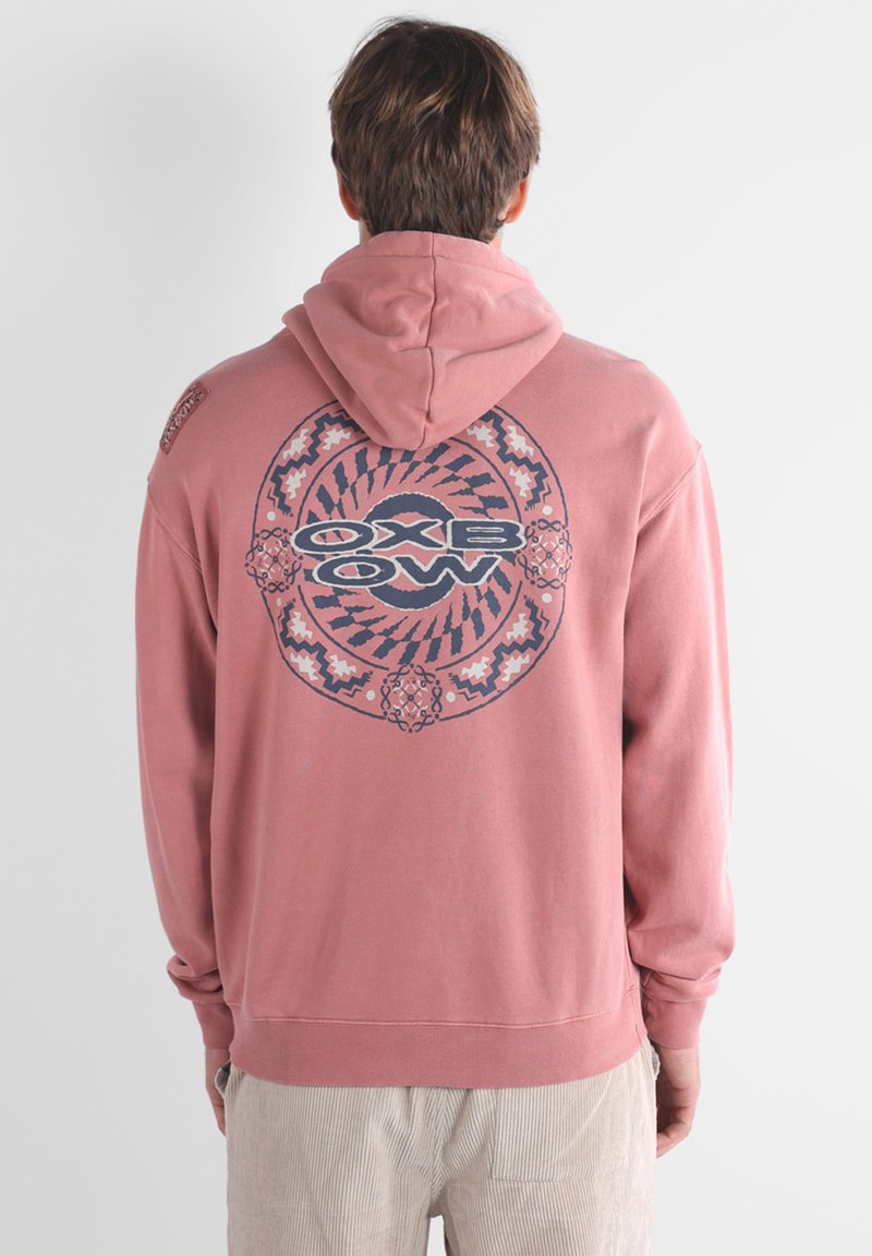 Felpa rosa con una grande grafica circolare blu scuro sul retro che presenta il testo "OXBOW" e motivi intricati. Tessuto di cotone, vestibilità comoda.