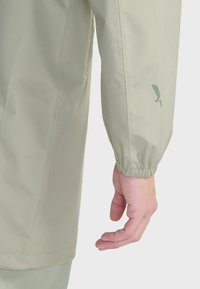 Svetlozelená bunda s elastickým manžetovým rukávom; hladká textúra; minimalistický dizajn; malý logotyp na manžete; mäkký, vodoodolný materiál.