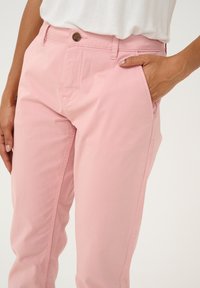 Light pink trousers with a smooth cotton texture, featuring a button closure and side pockets, partially depicted with a hand on the waistband.