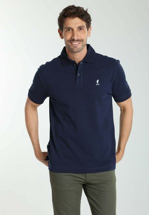 Homme souriant portant un polo bleu foncé et un pantalon vert olive, debout les mains derrière le dos sur un fond uni.