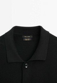 Polo en maille texturée noire avec col côtelé et fermeture à un seul bouton, avec une étiquette à l'intérieur de l'encolure.
