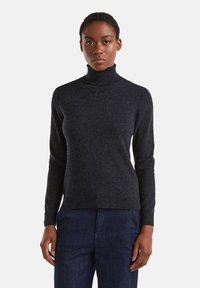 Femme portant un pull à col roulé gris foncé et un pantalon bleu foncé, se tenant devant un fond blanc uni.