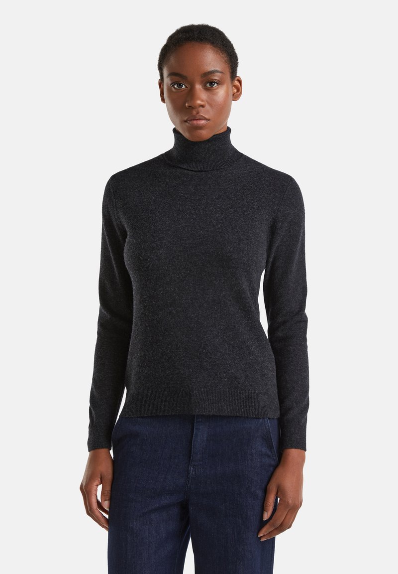 Femme portant un pull à col roulé gris foncé et un pantalon bleu foncé, se tenant devant un fond blanc uni.