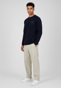 Sudadera azul marino de manga larga, cuello redondo y detalle sutil de logotipo. Combinada con pantalones beige claro y zapatillas blancas.