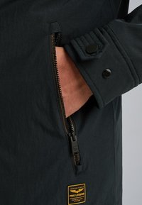 Donkergrijze jas met een ritszak met bronzen ritsdetails. Inclusief een gestructureerde schouder en een gele merktag.