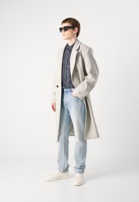 Cappotto grigio in lana sopra una camicia a fantasie scure, jeans azzurri chiari e sneakers bianche. Modello con occhiali da sole neri in posizione standing.