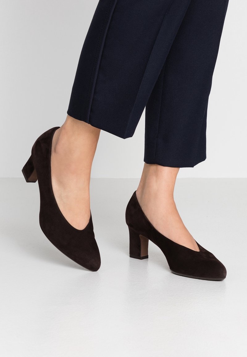 Femme portant des chaussures en daim noir à talon bas bloc associées à un pantalon court foncé sur un sol clair.